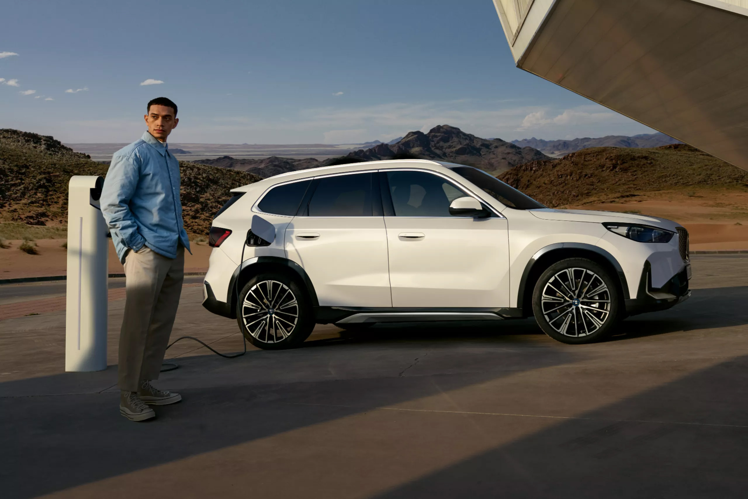 Auf diesem Bild ist ein Mann zu sehen, der neben einem weißen Elektroauto steht, das an einer Ladestation in einer bergigen Wüstenlandschaft aufgeladen wird.