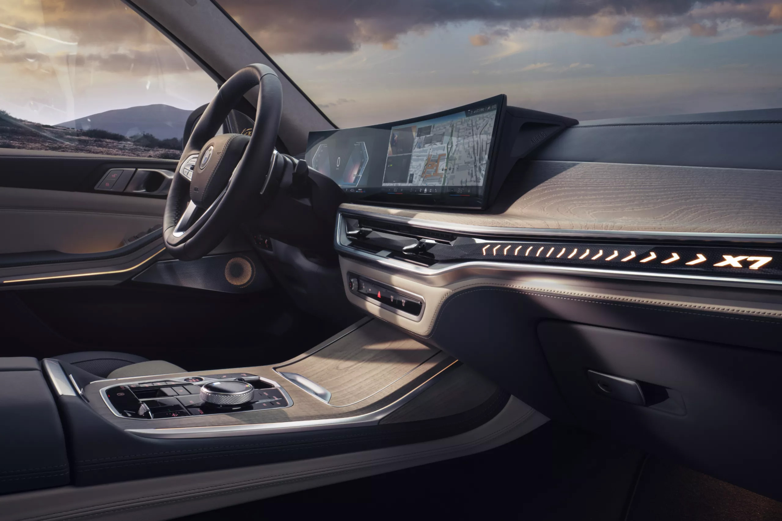 Das Bild zeigt den modernen und luxuriösen Innenraum eines BMW X7 bei Sonnenuntergang, mit dem Lenkrad, einem breiten Curved Display und einer beleuchteten 'X7'-Schrift im Armaturenbrett.