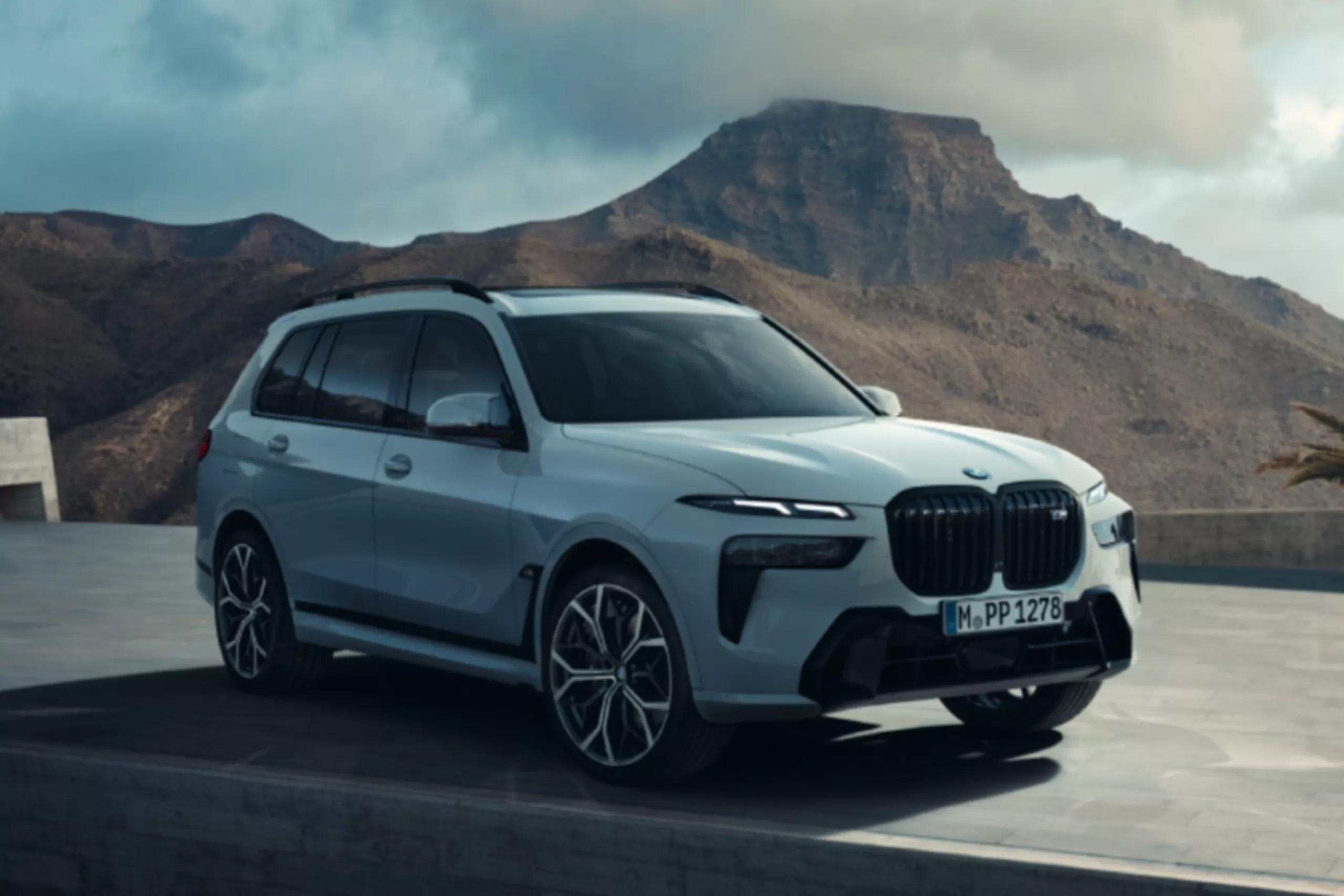 Das Bild zeigt einen hellen BMW X7 SUV, der vor einer felsigen Berglandschaft unter einem bewölkten Himmel steht.