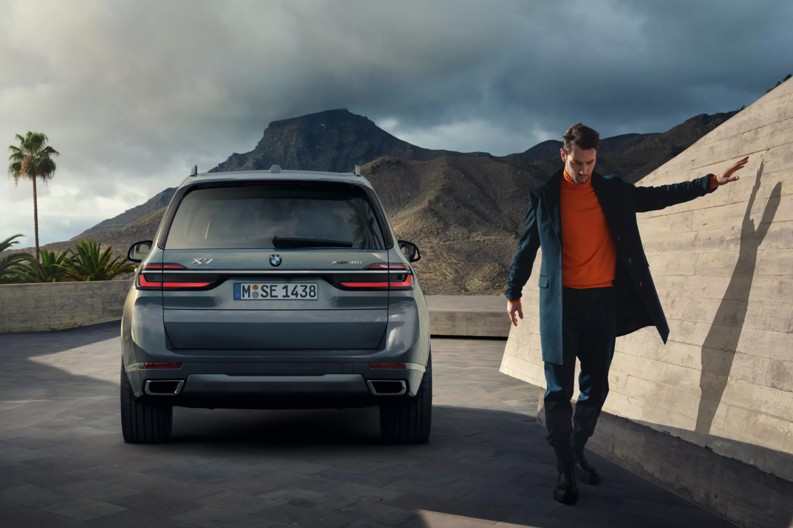 Auf diesem Bild ist ein grauer BMW X7 SUV zu sehen, neben dem ein Mann vor einer Kulisse aus Bergen und einer schrägen Betonwand entlanggeht.