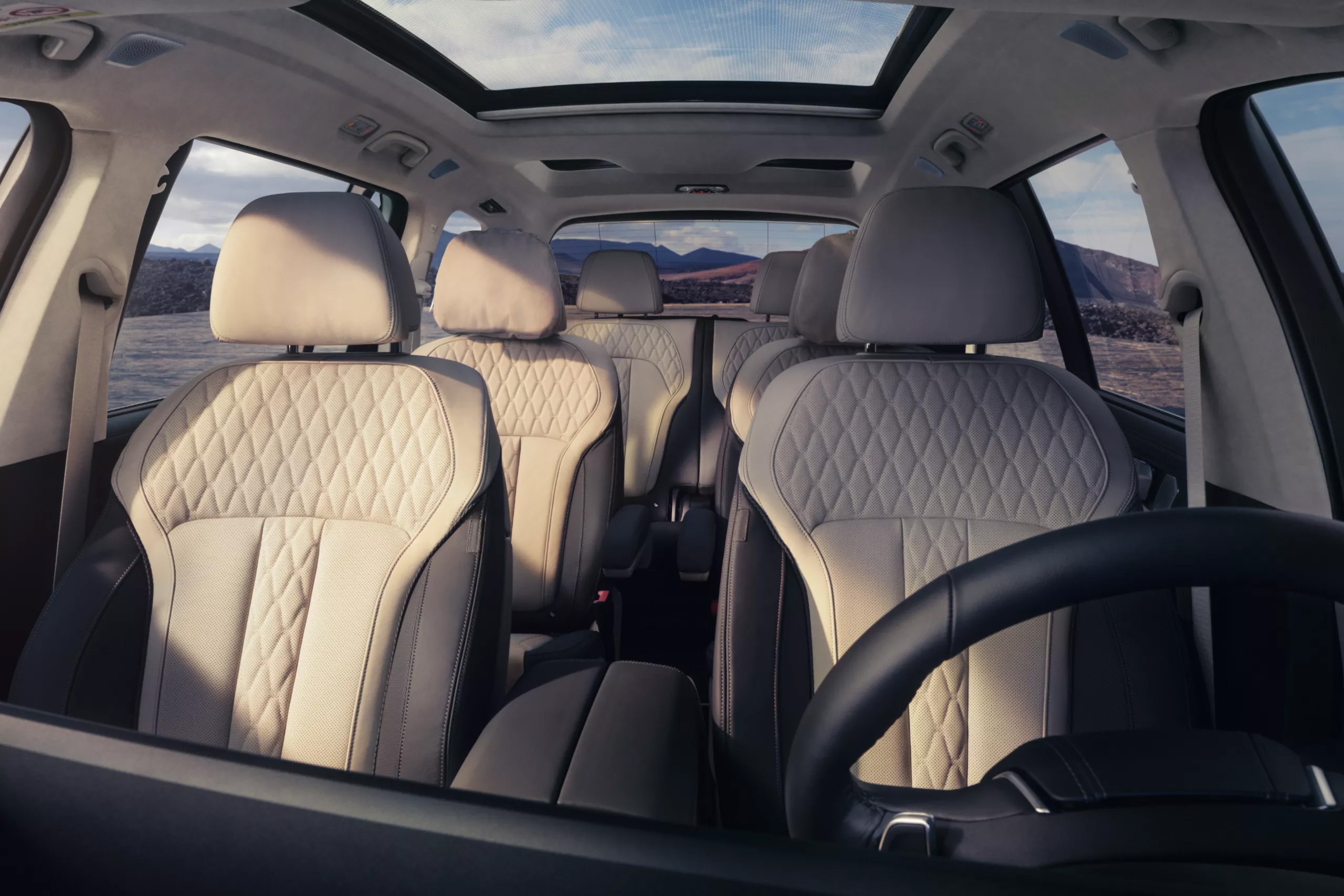 Auf diesem Bild ist der geräumige Innenraum eines modernen Autos mit mehreren Reihen von zweifarbigen, gesteppten Sitzen und einem großen Panoramadach zu sehen, das den Blick auf eine weite Landschaft unter blauem Himmel freigibt.