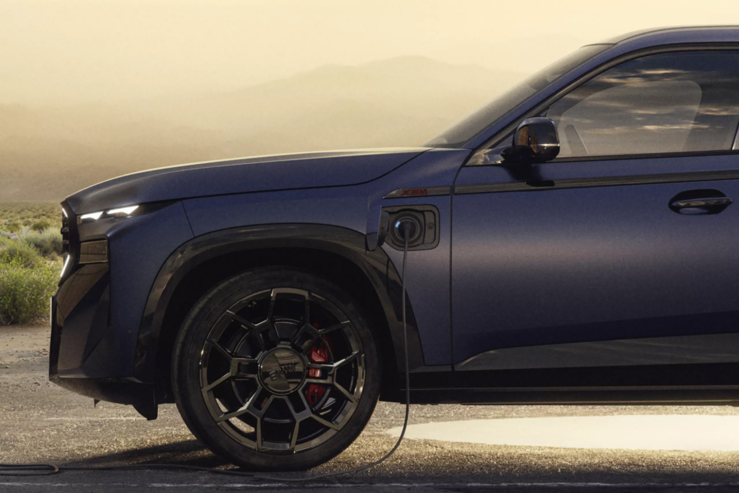 Auf diesem Bild ist ein dunkelblauer BMW XM zu sehen, der in einer wüstenähnlichen Landschaft über ein Ladekabel aufgeladen wird.