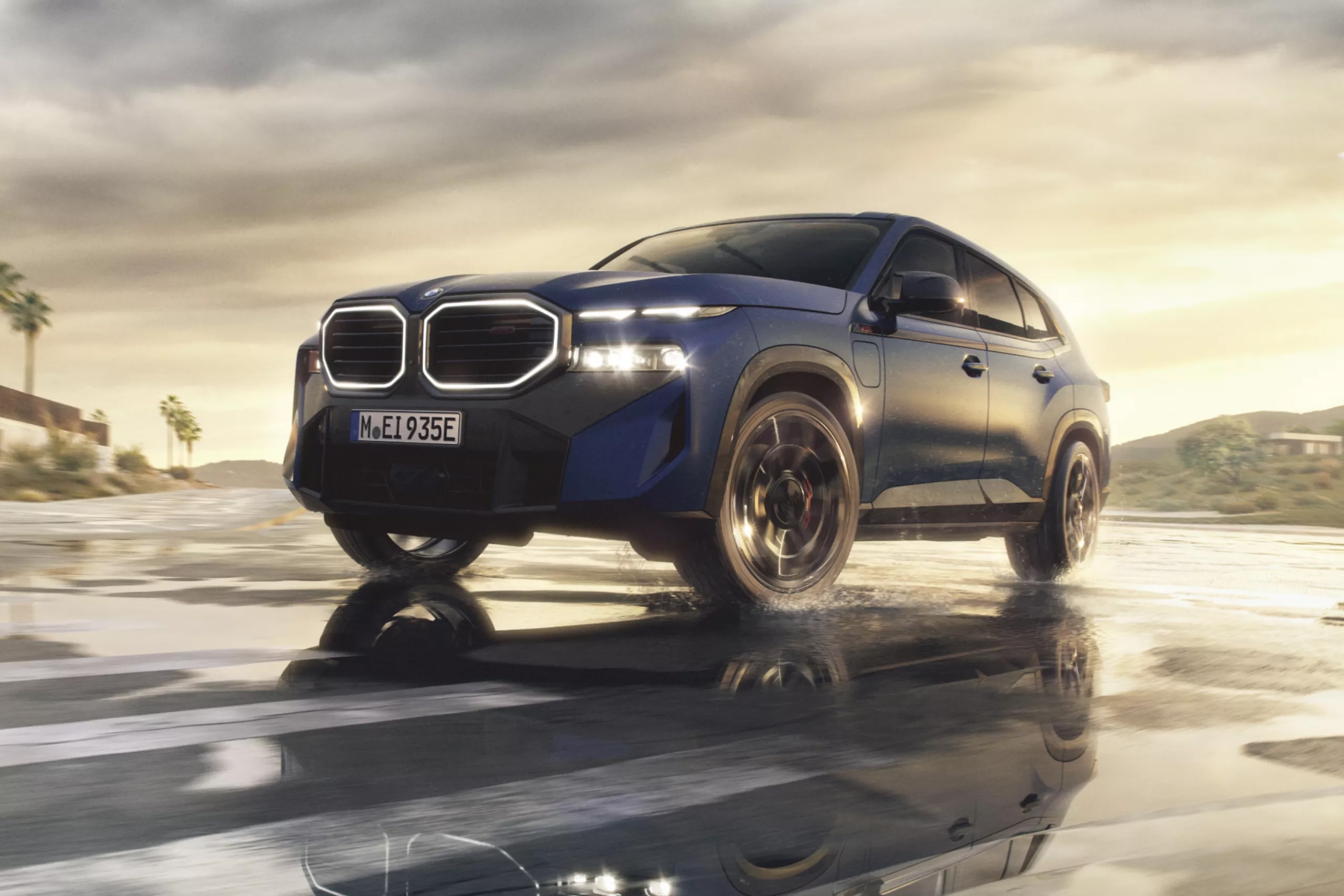 Auf diesem Bild ist ein blauer BMW SUV zu sehen, der dynamisch auf einer nassen Straße fährt, dabei Wasser aufwirbelt und seine Lichter bei dämmerigem Licht leuchten lässt.