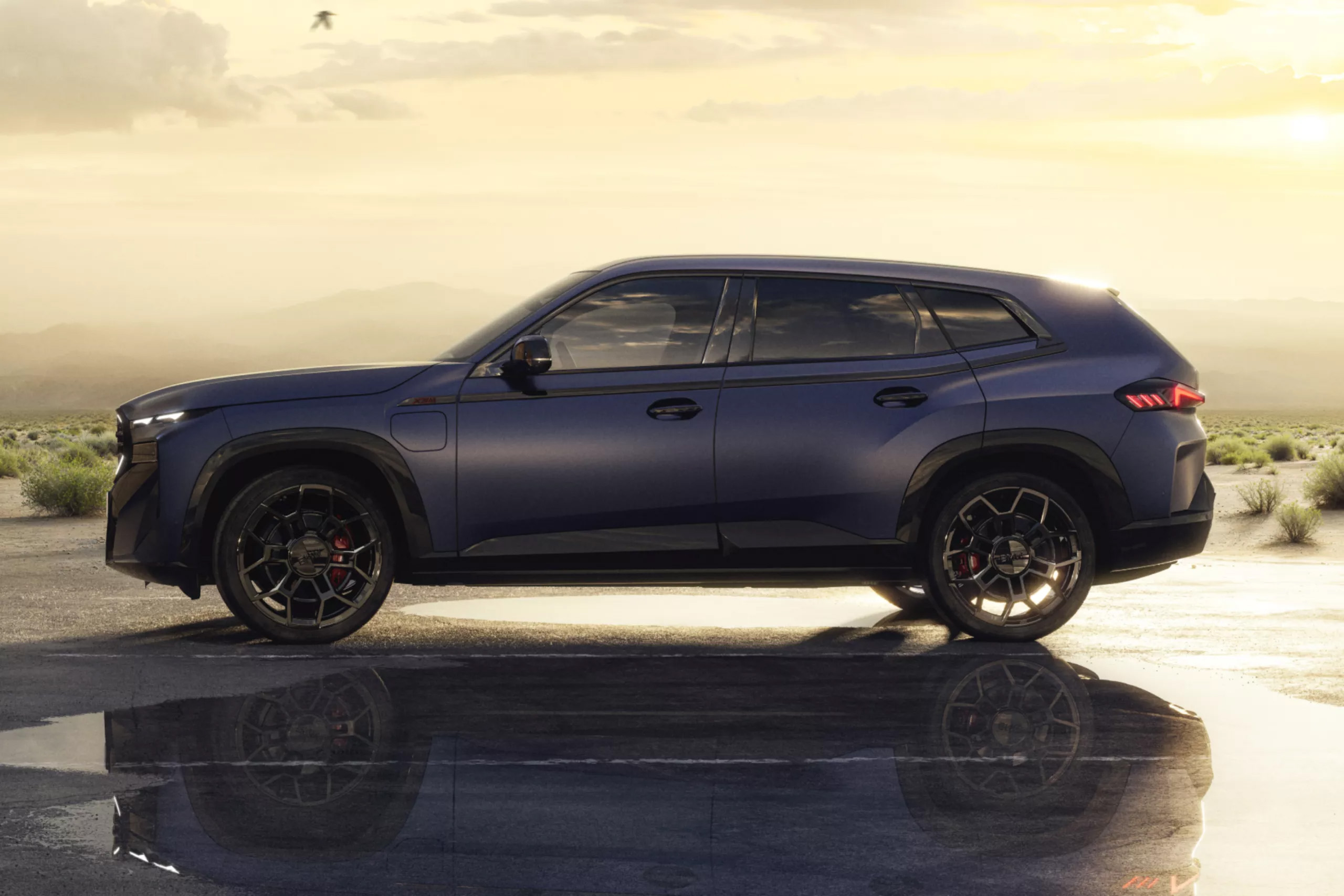 Auf diesem Bild ist ein dunkelblaues SUV in einer kargen, bergigen Landschaft bei Sonnenuntergang abgebildet, das sich in einer Pfütze auf dem Boden spiegelt.